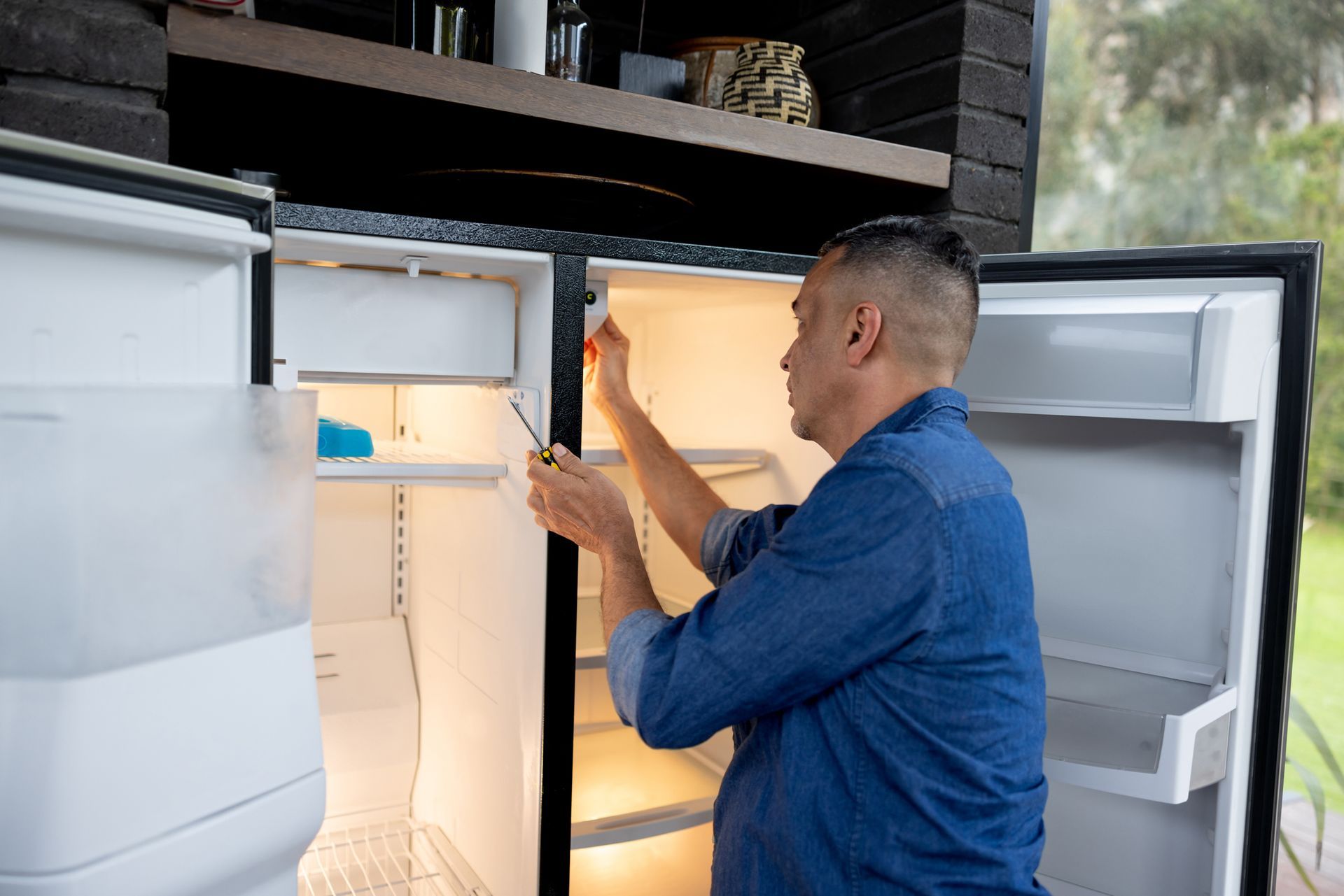A man wearing a blue denim shirt is fixing an appliance, demonstrating refrigerator repair skills.