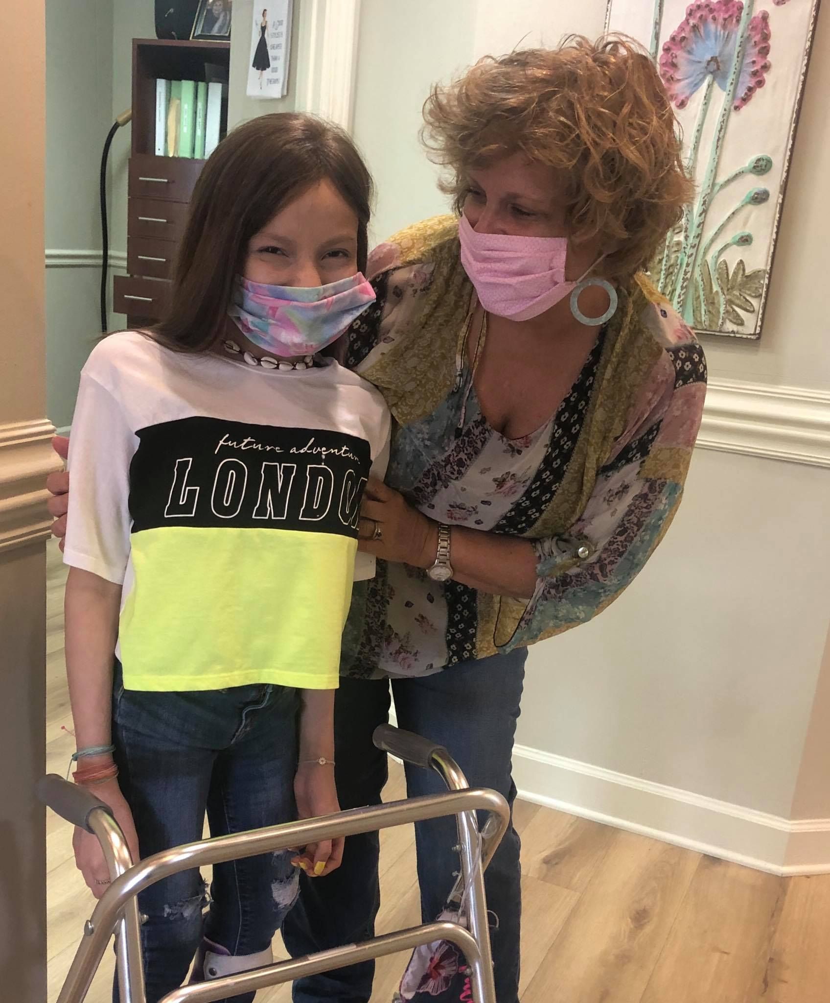 A young girl and woman wearing masks, posing with a walker indoors. The girl smiles.