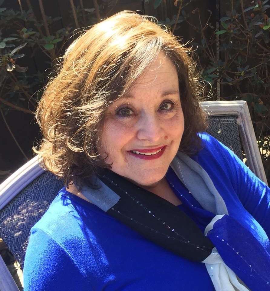 Woman with brown hair smiling, wearing a blue top, black and gray scarf, sitting outside.