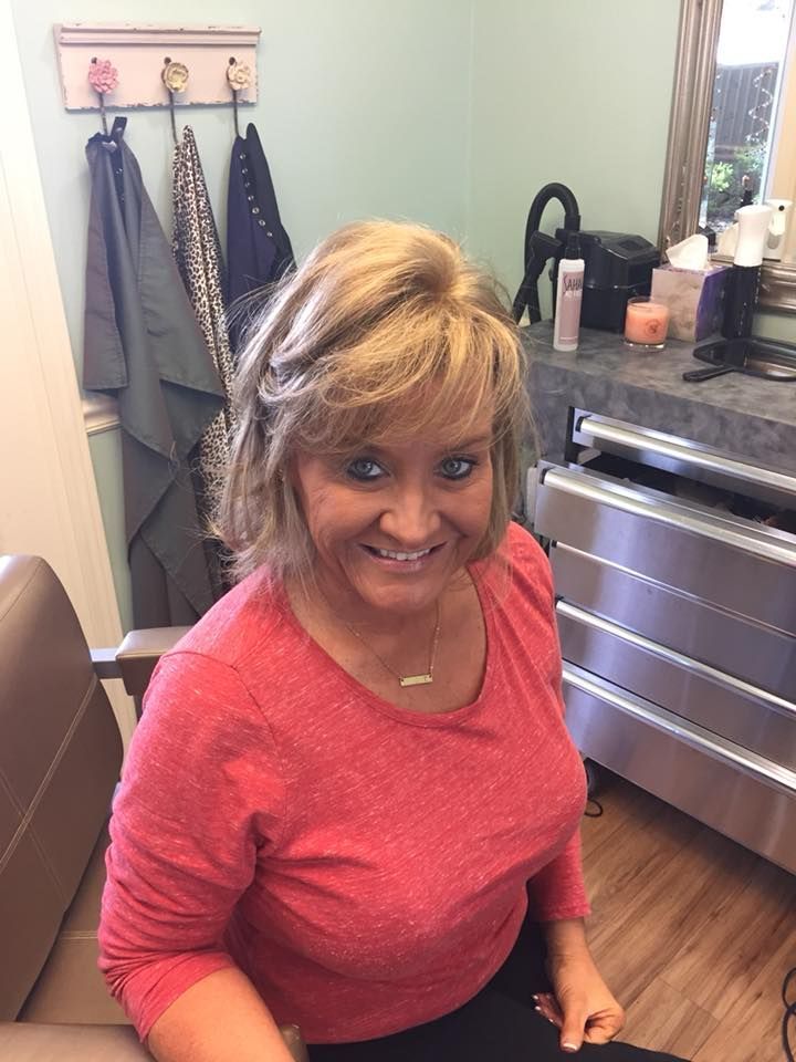 Woman with blonde hair in a red shirt smiles at the camera; seated in a salon with light green wall and stainless steel counter.