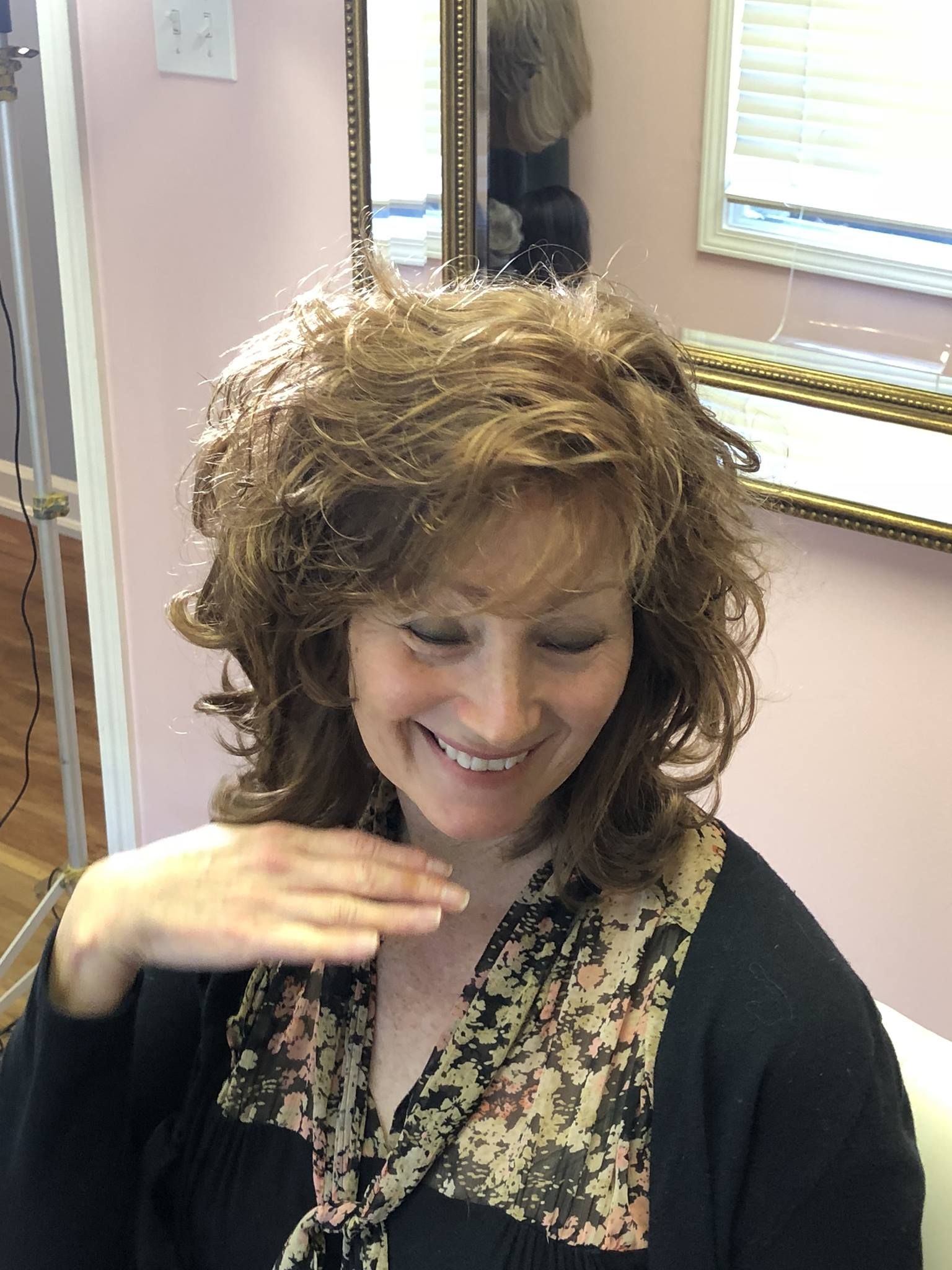 Woman with wavy brown hair smiling, hand near her face, in a salon setting.