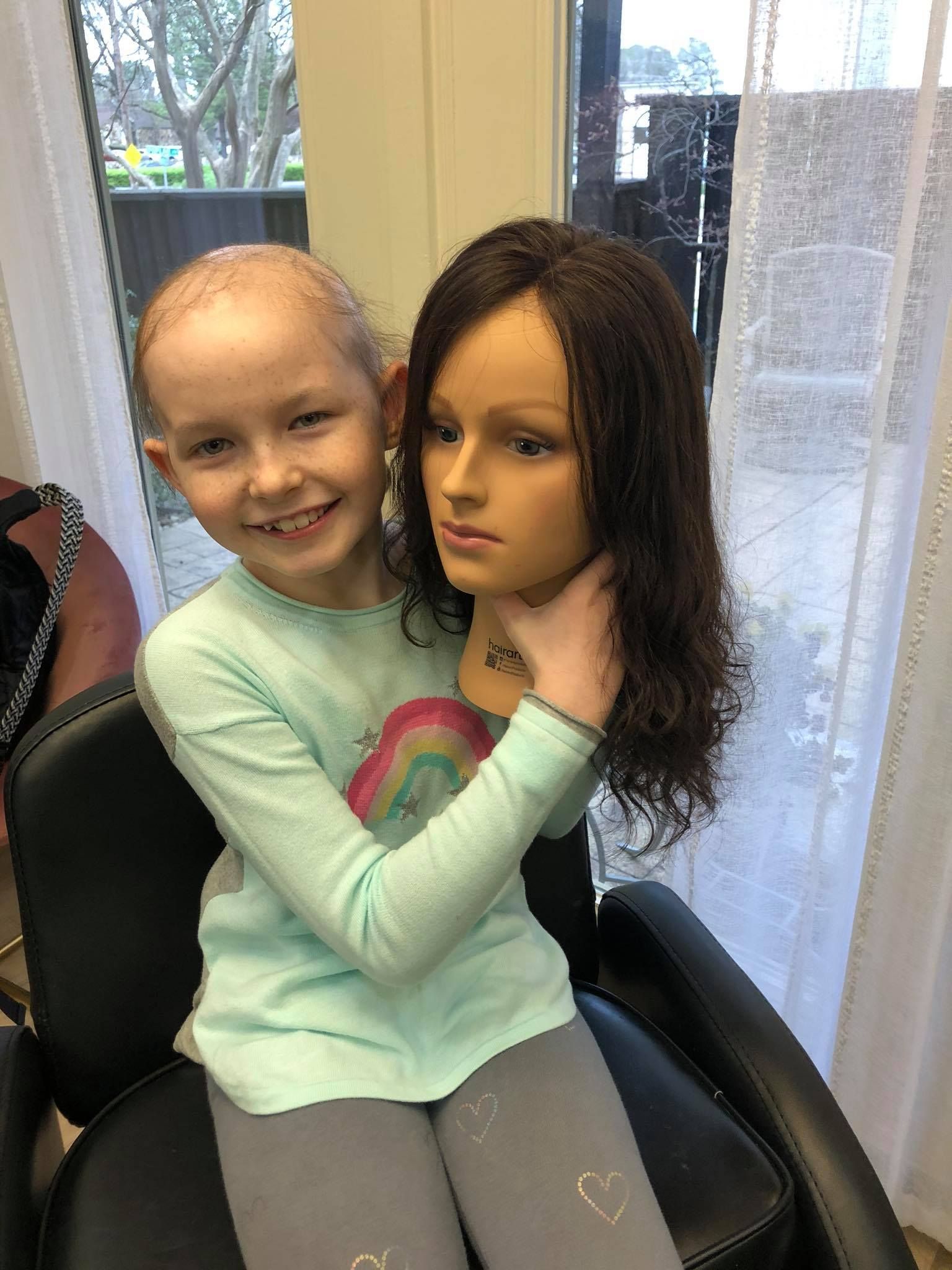 Smiling child holding a mannequin head with a wig on, sitting in a salon chair.