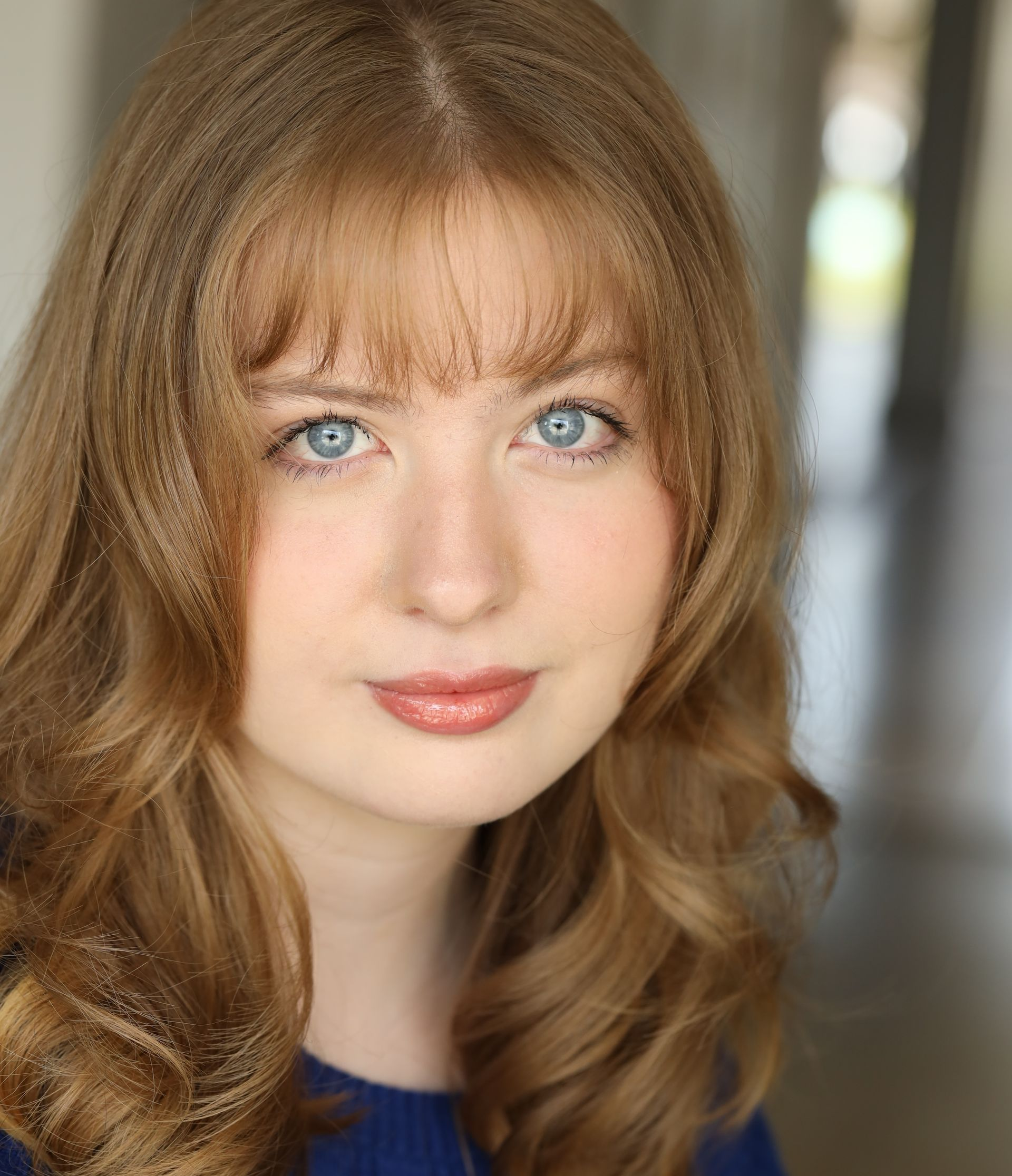 Woman with light brown hair, blue eyes, and bangs, looking at the camera. She wears a blue top.