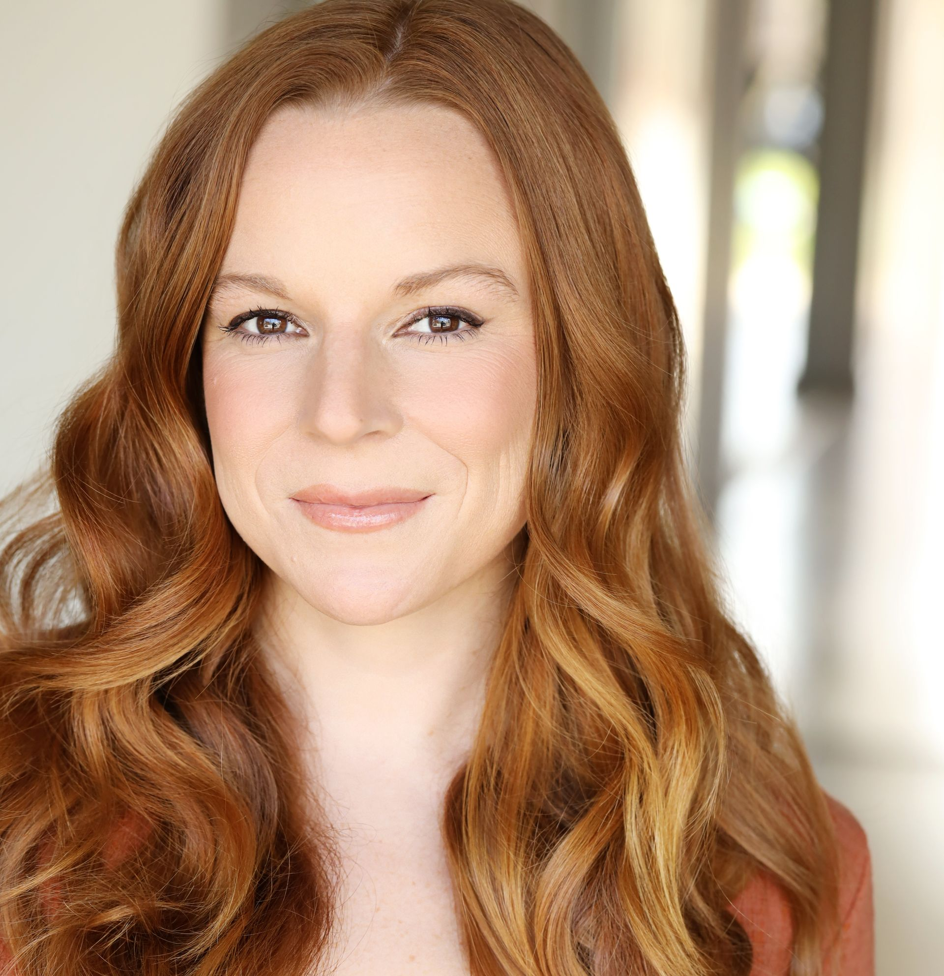 Woman with long, wavy auburn hair smiles, wearing a brown jacket, indoors, with a blurred background.