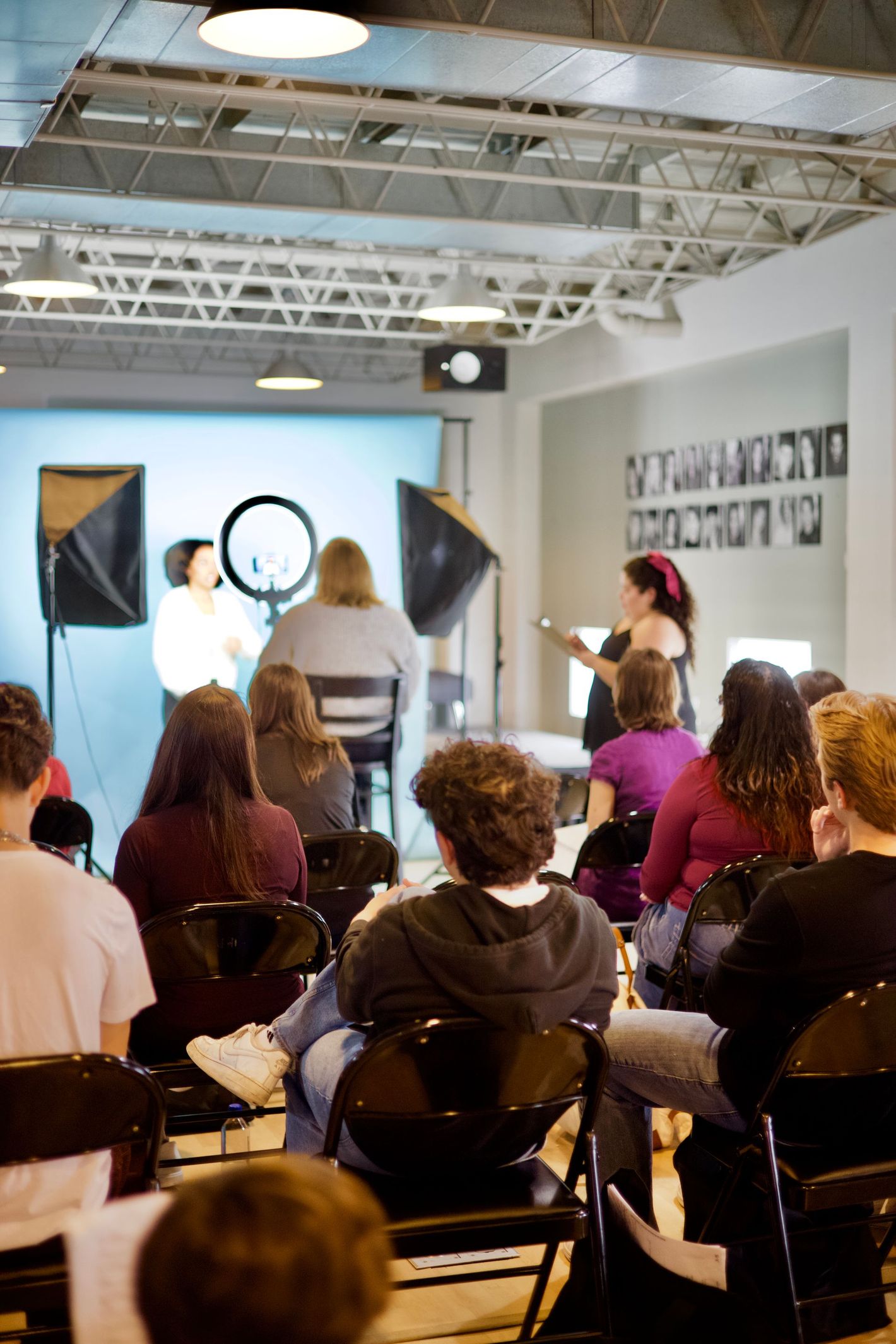 A group of people watch a demonstration in a studio with lights. A person in the background is being photographed.