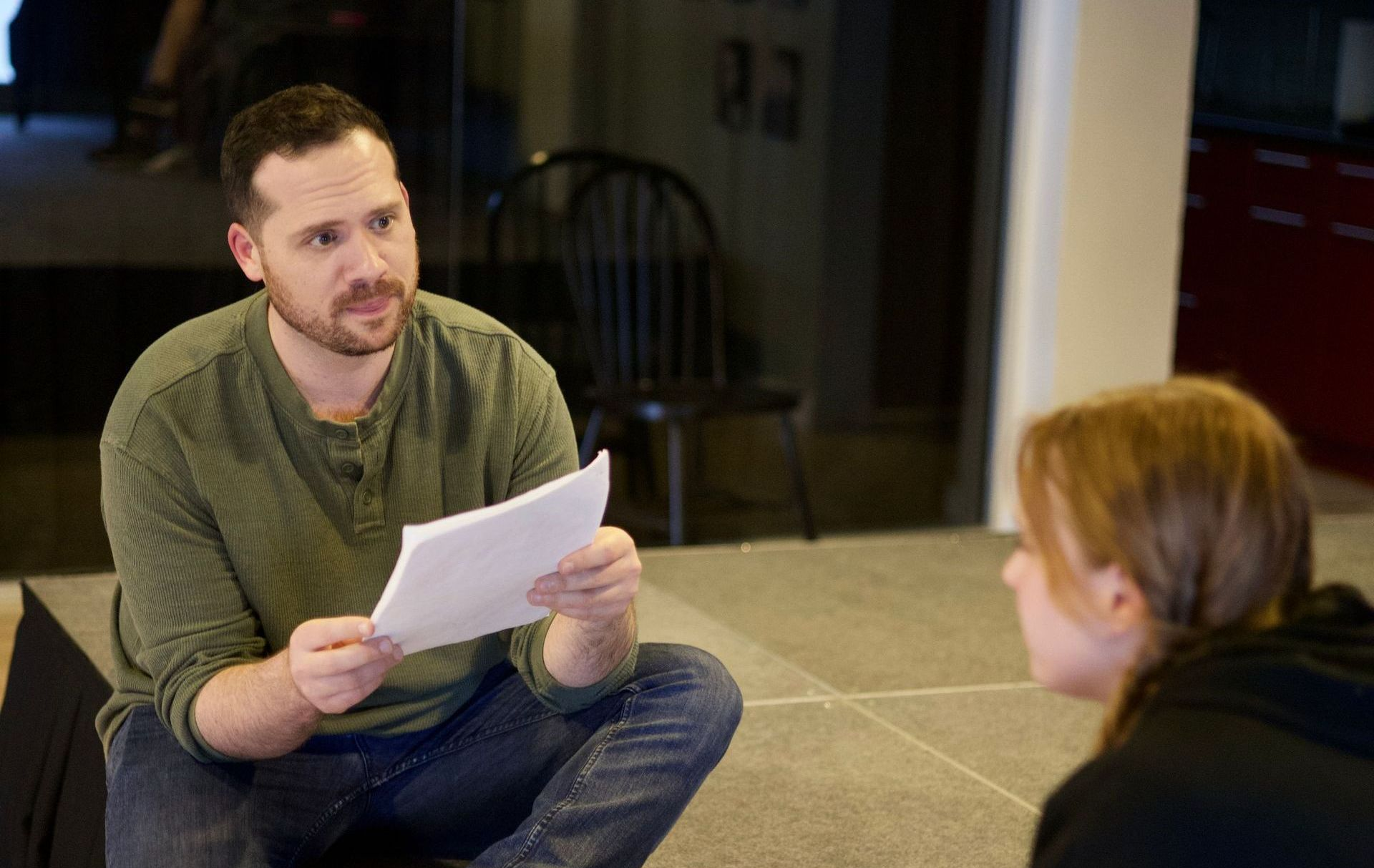 Man holding paper, talking to a woman. They are indoors. Man is wearing green shirt.