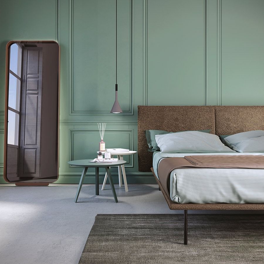 Camera da letto color verde pastello con letto, comodino, specchio e luce naturale soffusa.
