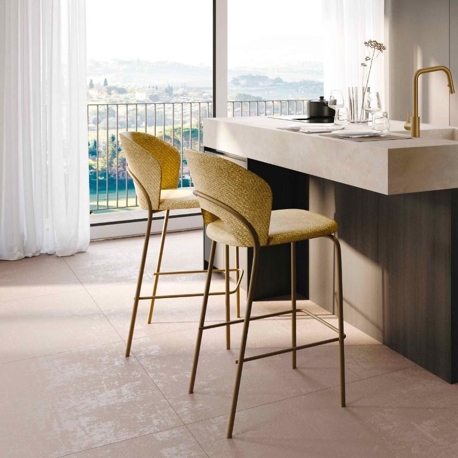 Cucina moderna con due sgabelli da bar dorati accanto a un'isola in marmo e ampie finestre che si affacciano su un balcone.