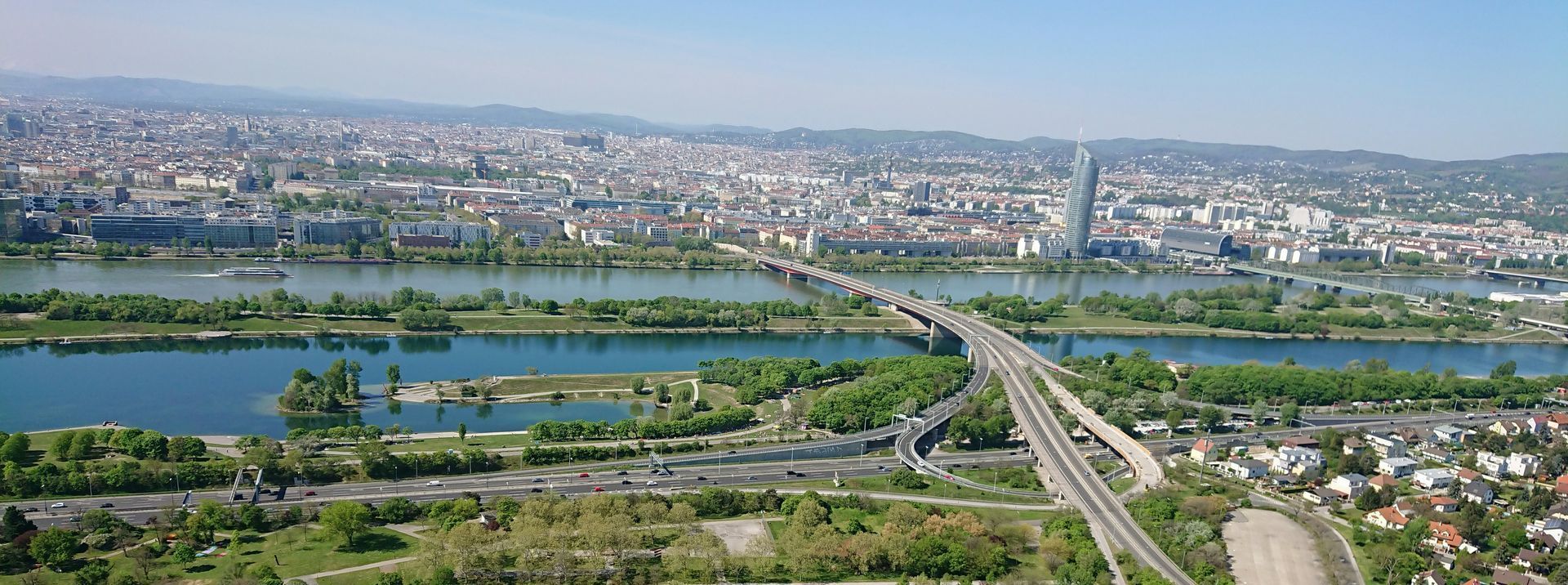 Aerial view of the Danube River in Vienna, showing bridges, green parks, and modern buildings stretching across the city.