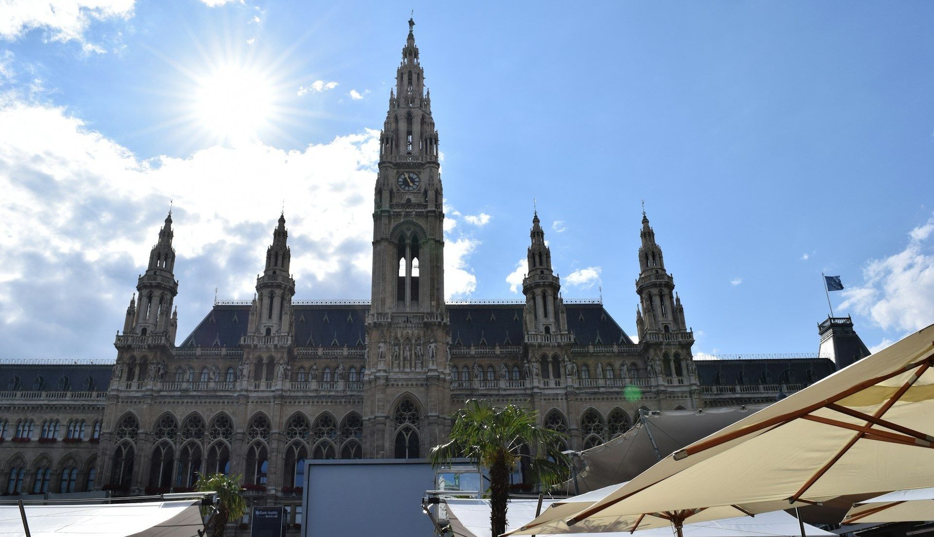 Front view of Vienna City Hall at Rathausplatz under a bright blue sky, with people and market stalls in the foreground.
