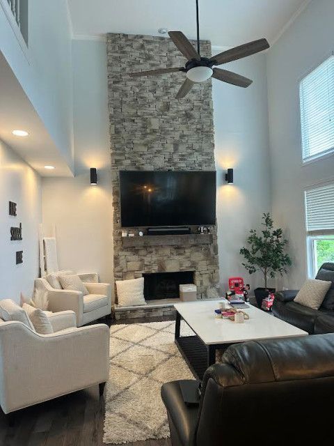 Bright living room with stone fireplace, wall-mounted TV, high ceiling, beige chairs, and dark leather sofa