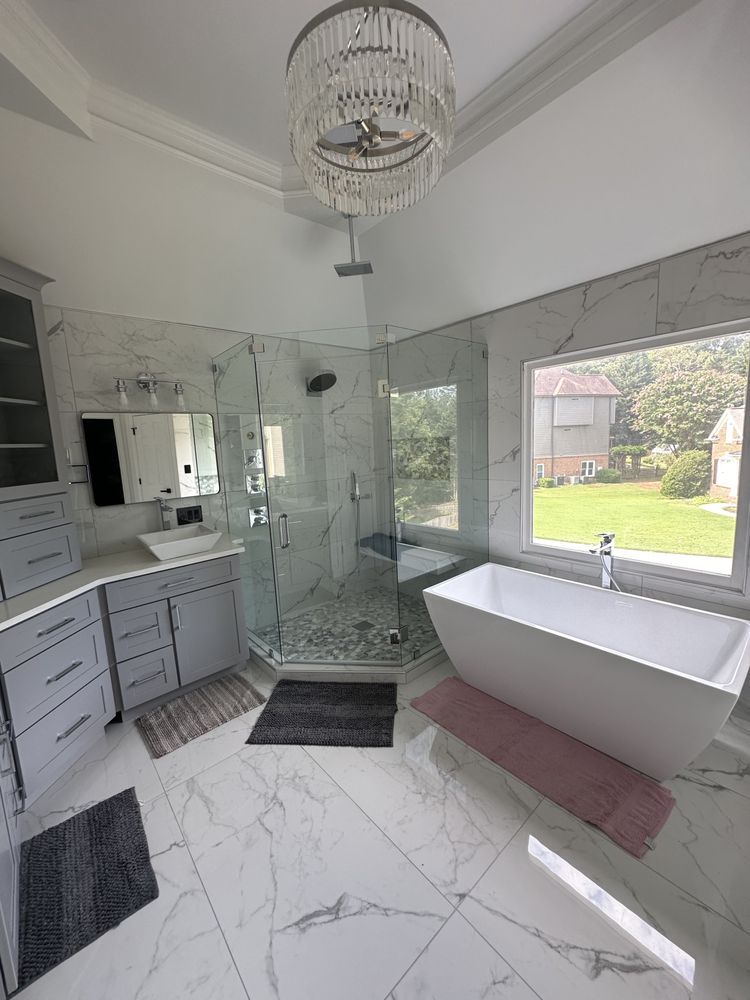 Modern marble bathroom with double vanity, glass shower, chandelier, and large window overlooking a yard