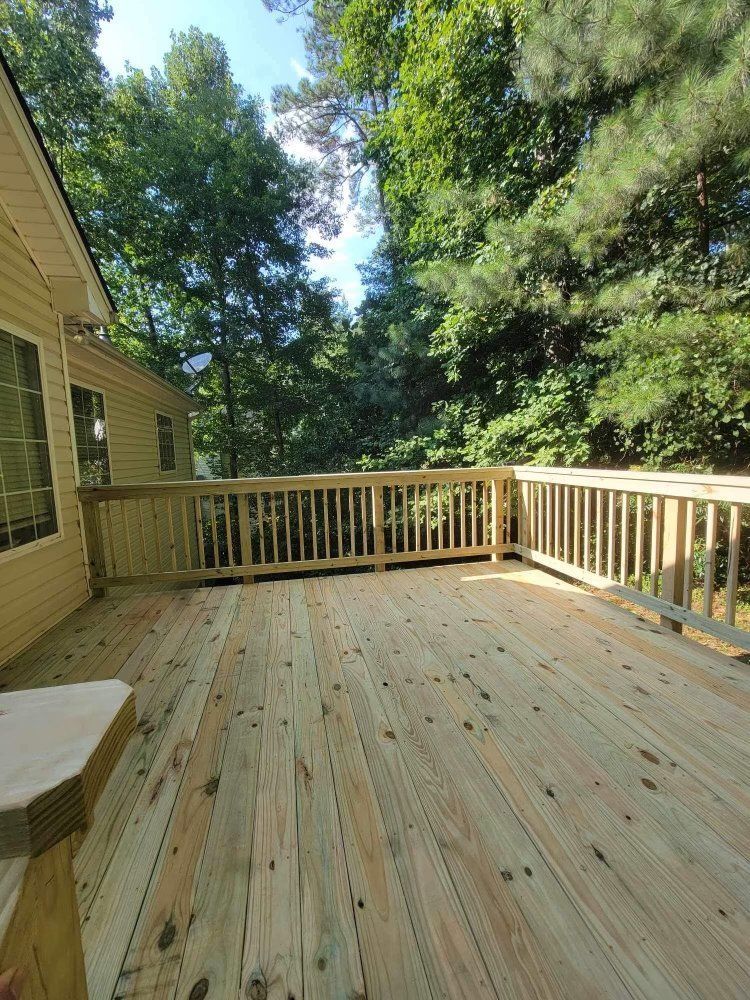 Wooden deck with railing beside a house, surrounded by tall trees and greenery under bright sunlight.