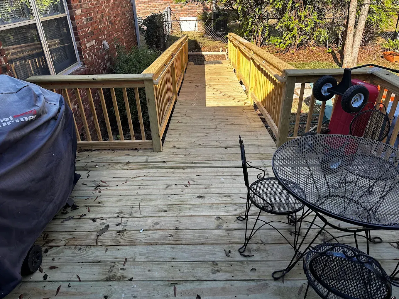 Wooden deck walkway with yellow railings, patio table, grill, and a backyard garden beyond.