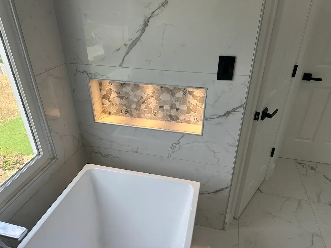 Modern bathroom with freestanding tub, marble walls, lit recessed niche, and window on the left