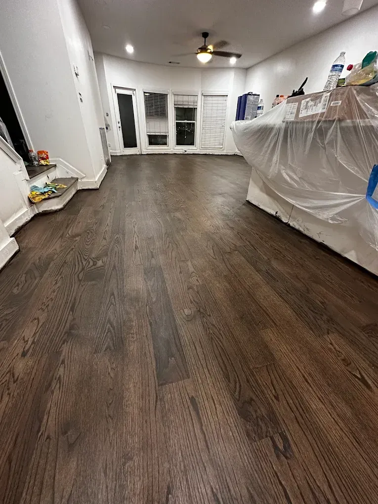 Empty room with dark wood floor, white walls, plastic-covered items, and ceiling fan near glass doors