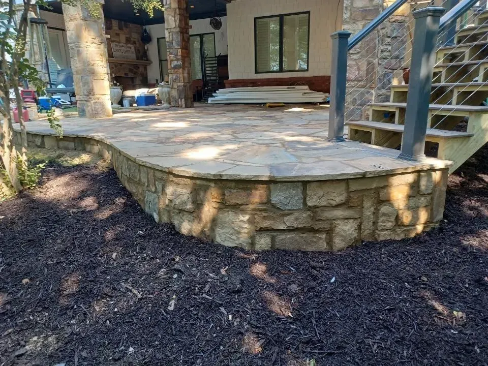 Stone patio with curved retaining wall and stairs leading to a house porch, surrounded by mulch and trees