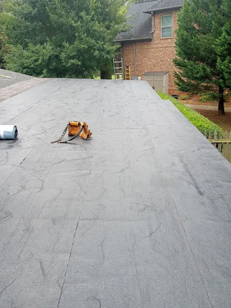 Workers on a flat roof applying roofing material near a brick house and trees