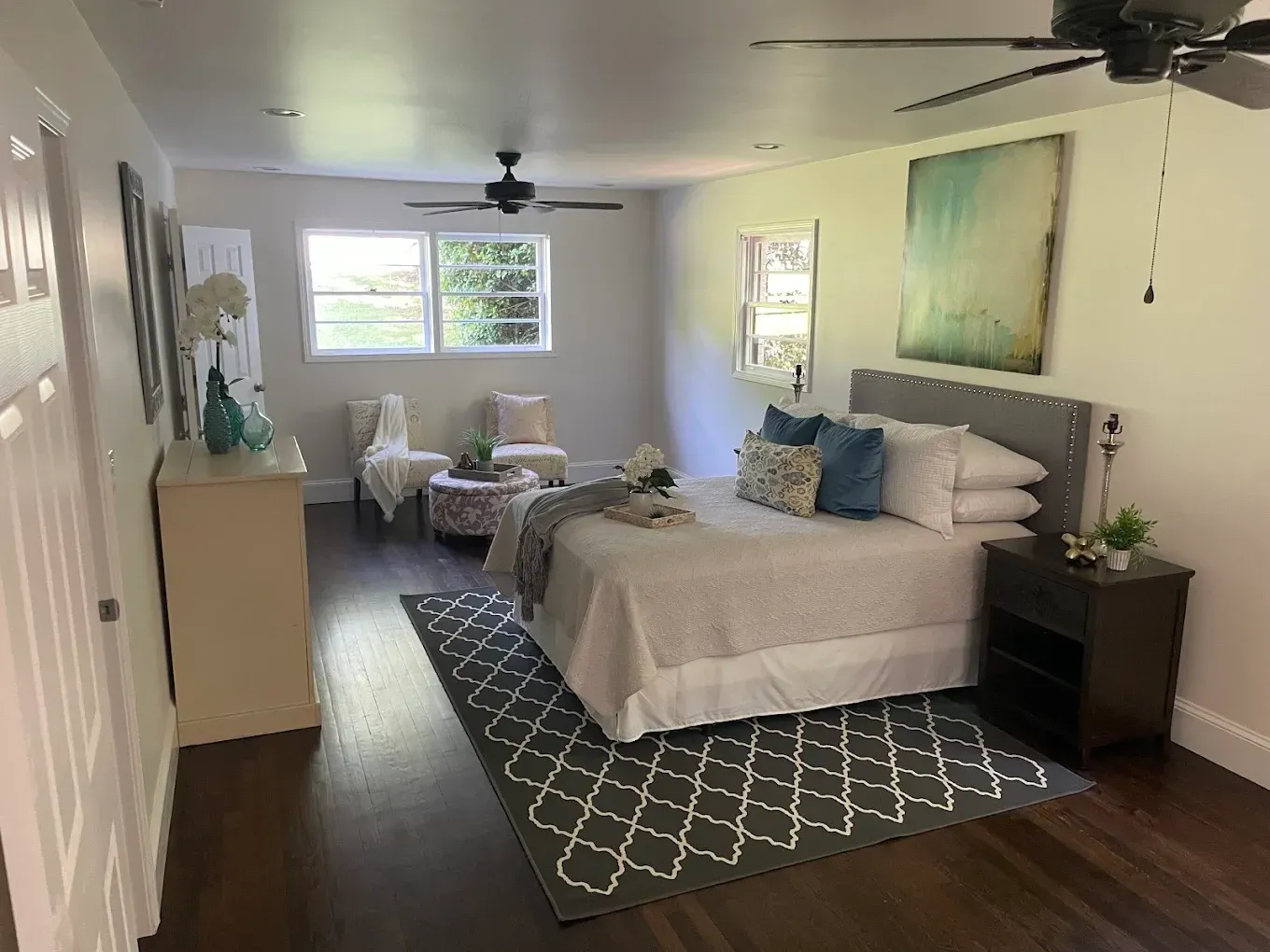 Bright bedroom with a bed, rug, nightstands, ceiling fan, and sitting area by the window.