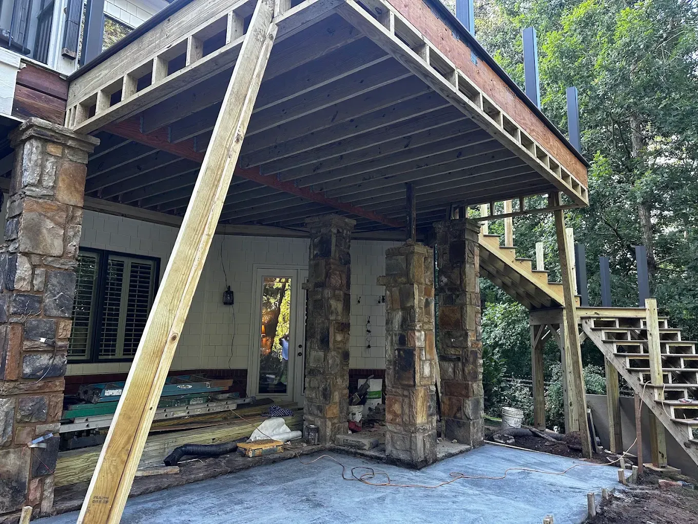 Partially built stone patio with wooden deck above, support beams, and stairs beside a forested home