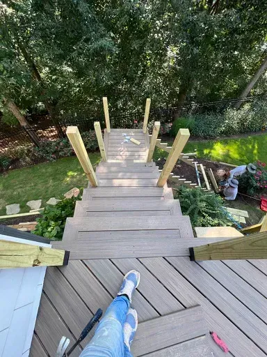 Person standing at top of a steep wooden staircase with railings, overlooking a wooded yard
