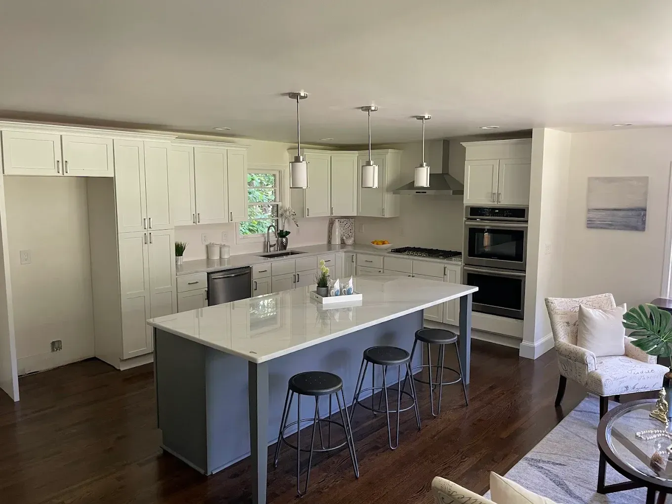 Modern kitchen with white cabinets, gray island, bar stools, and dark hardwood floors, opening to a seating area
