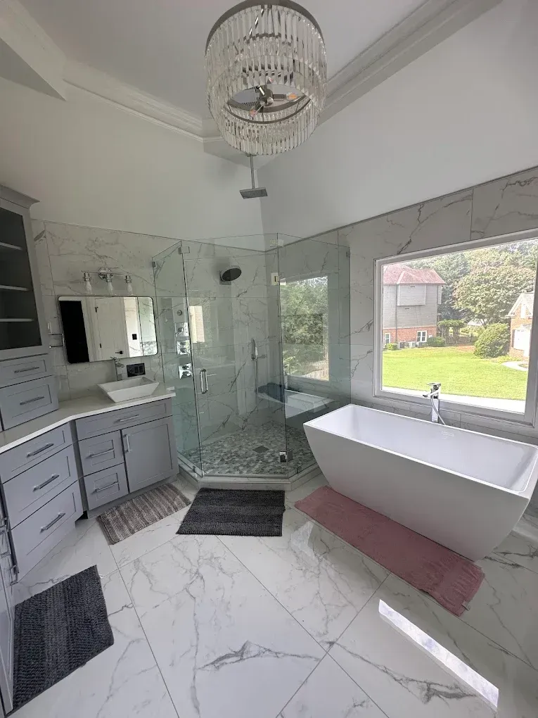 Modern marble bathroom with glass shower, freestanding tub, vanity, and chandelier, overlooking a green yard