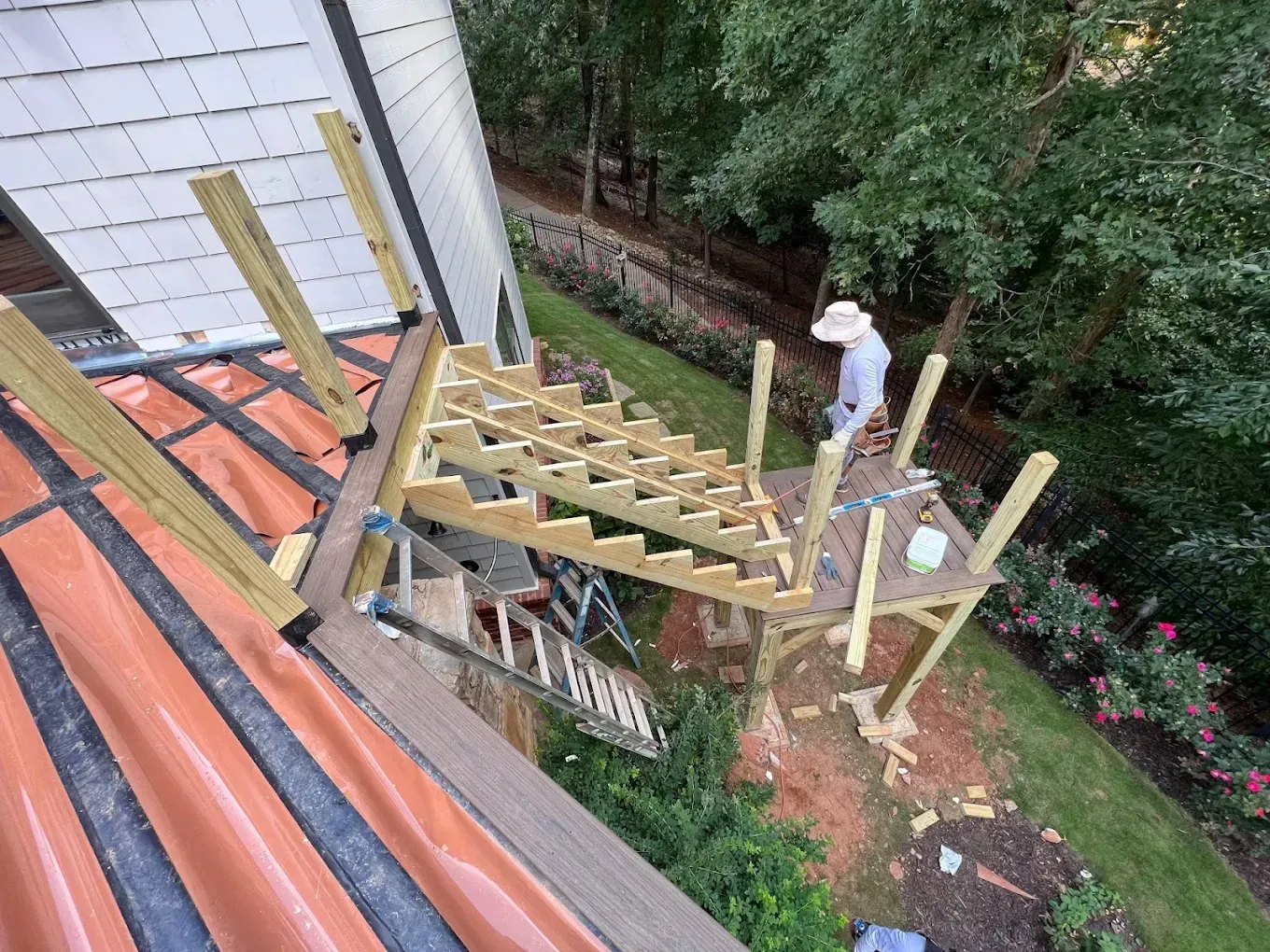 Deck framing under construction beside a house, with a worker in a white hat and backyard greenery.