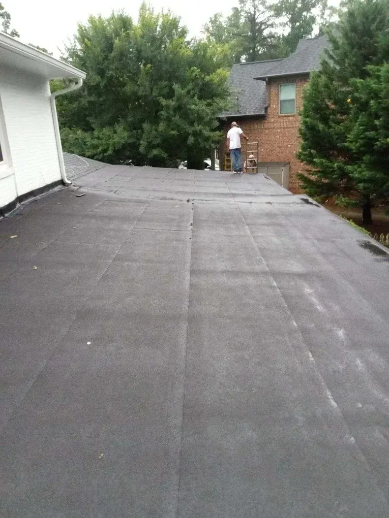 Flat dark roof between a white house and brick building, with trees in the background.