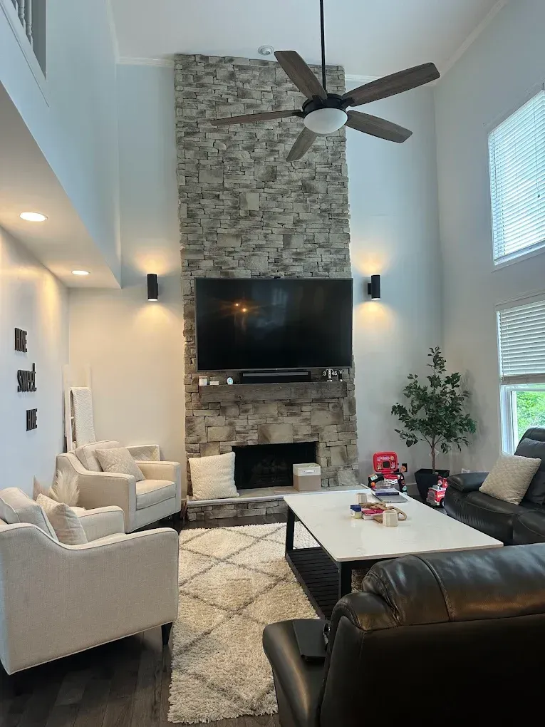 Bright living room with high ceiling, stone fireplace, TV, white chairs, and black leather sofa