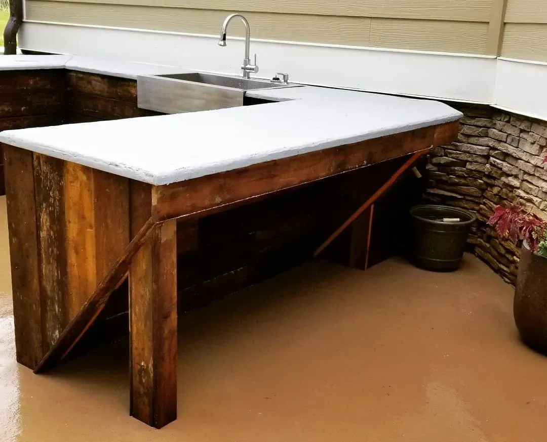 Outdoor rustic sink table with white countertop and wooden legs beside a stone wall and faucet