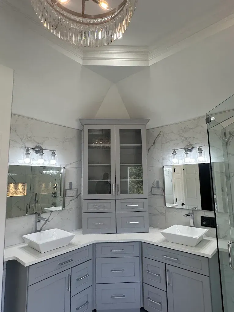 Elegant bathroom vanity with gray cabinets, marble countertops, mirrors, and a glass shower door