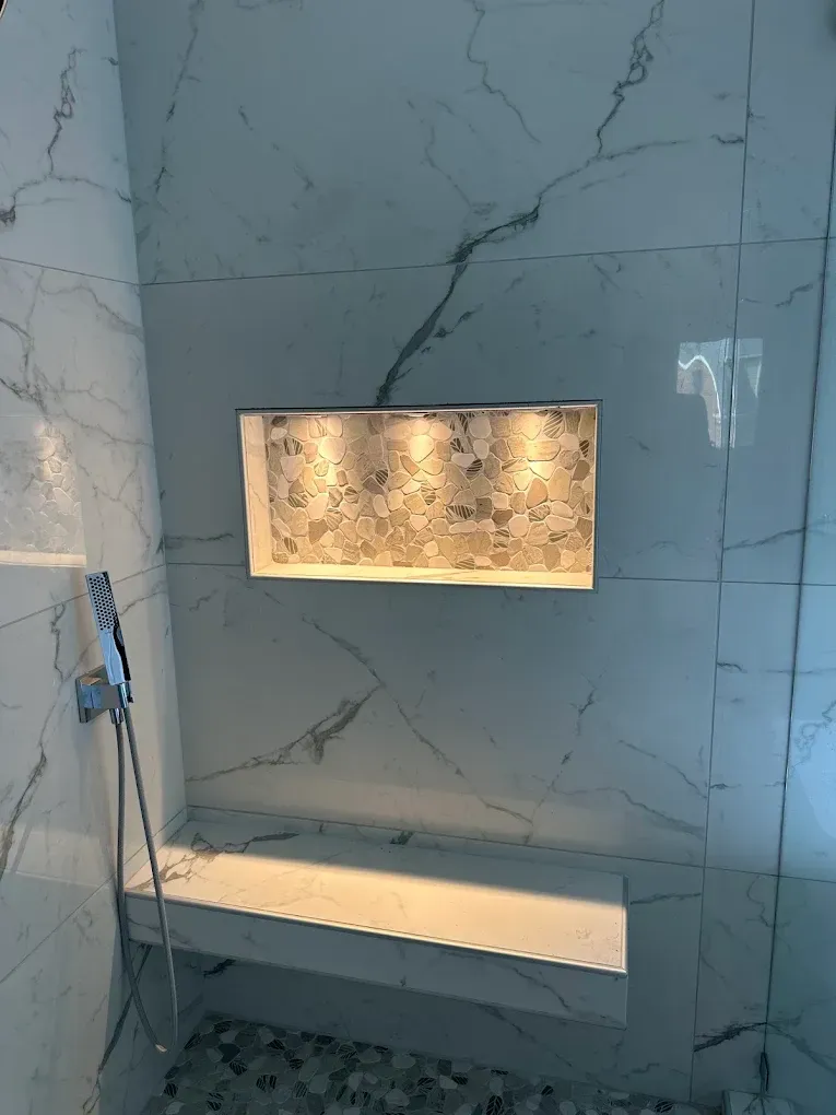 Marble-tiled shower with recessed lit shelf and built-in bench, featuring a hand-held showerhead on the left.