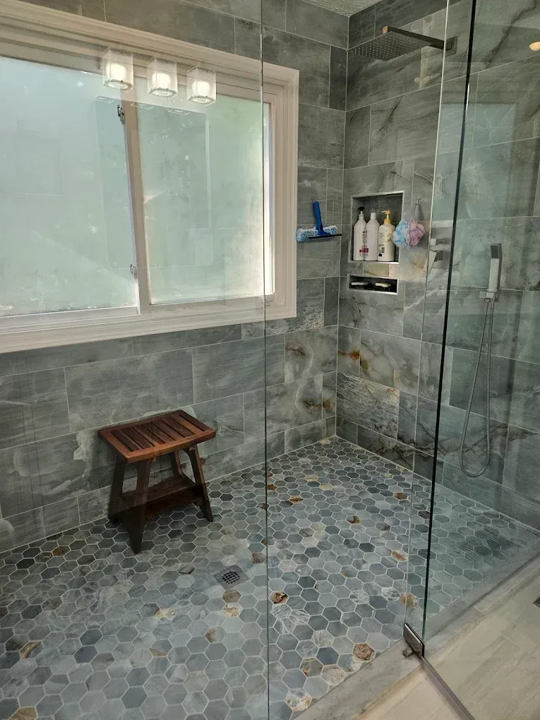 Gray tiled walk-in shower with glass door, window, built-in shelf, and small wooden stool.