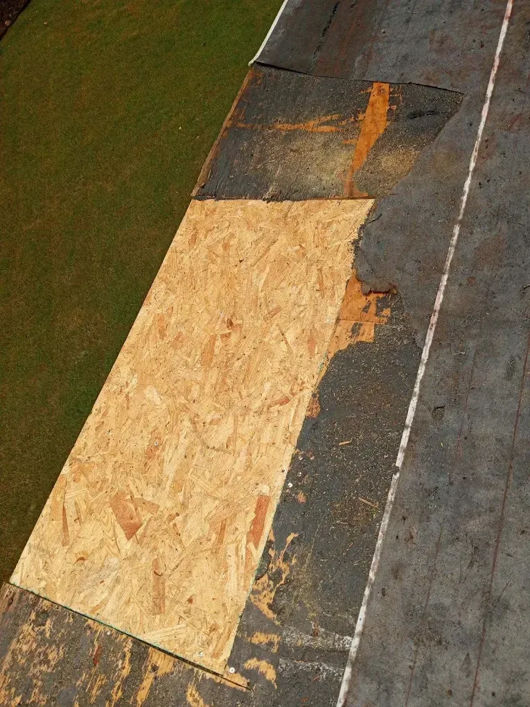 Damaged rooftop with a large plywood patch over missing shingles and surrounding dark roofing material
