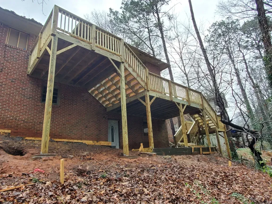 Wooden deck and stairs on a brick house, viewed from below on a wooded hillside with fallen leaves.