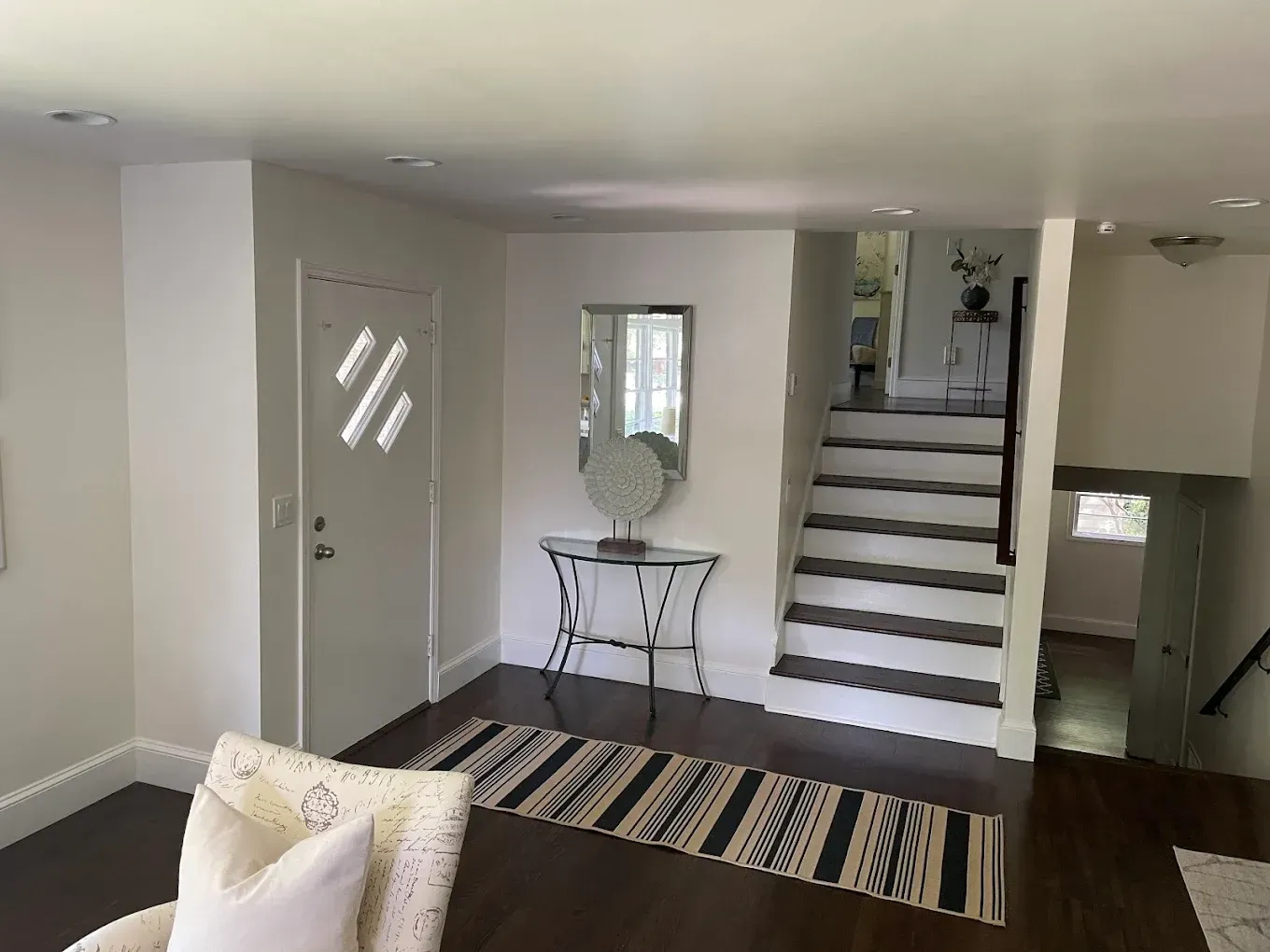 Bright entryway with staircase, striped rug, console table, and adjacent hallway and living room.