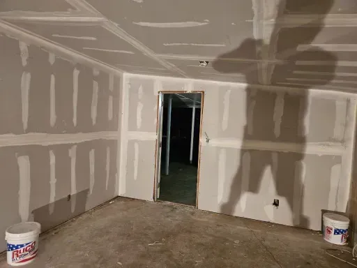 Unfinished drywall room with primer patches, buckets on the floor, and a doorway to a dark hallway.