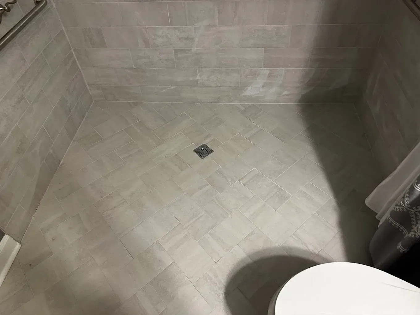 Tiled shower floor with a small square drain and a white toilet seat in the corner
