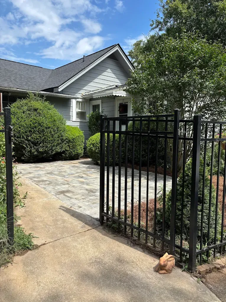 Black metal gate beside a stone walkway leading to a white house with shrubs and a tree