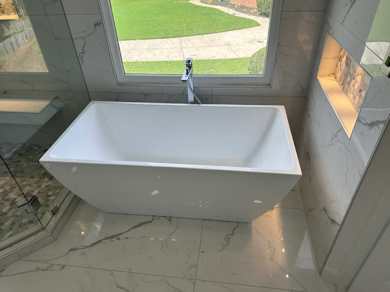 Freestanding white bathtub in a tiled bathroom with a large window and faucet above it
