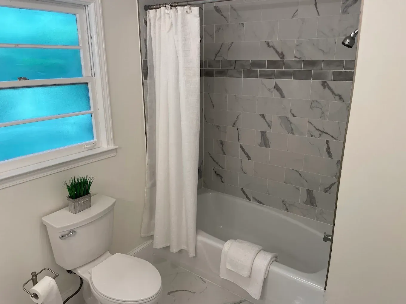 Bright bathroom with toilet, bathtub, window, and white shower curtain; small plant on the toilet tank.