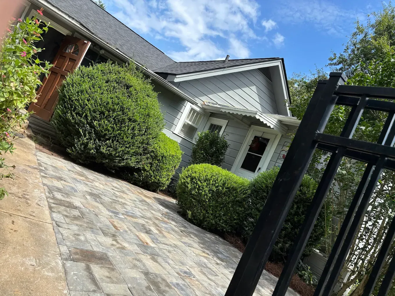 Tilted view of a house entrance with stone patio, trimmed green bushes, and a black gate under a blue sky