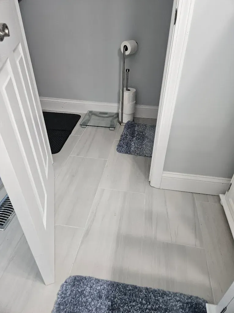 Narrow bathroom hallway with white doors, gray walls, and blue rugs on light wood floor.