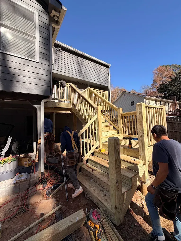 Workers installing a wooden deck staircase beside a gray house on a sunny day