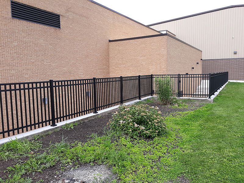 Black metal fence alongside a brick and tan building, enclosing a small garden.