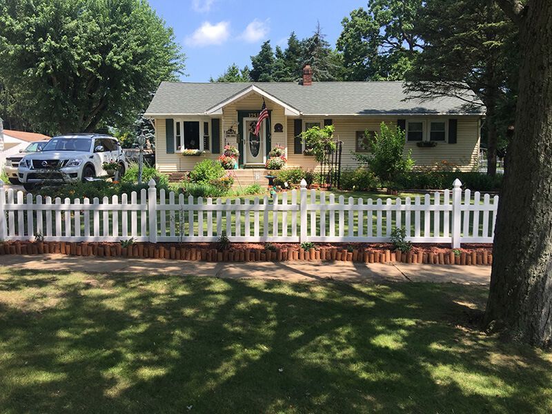 Cottage-style yellow house with white picket fence, brick base, and green lawn on a sunny day.