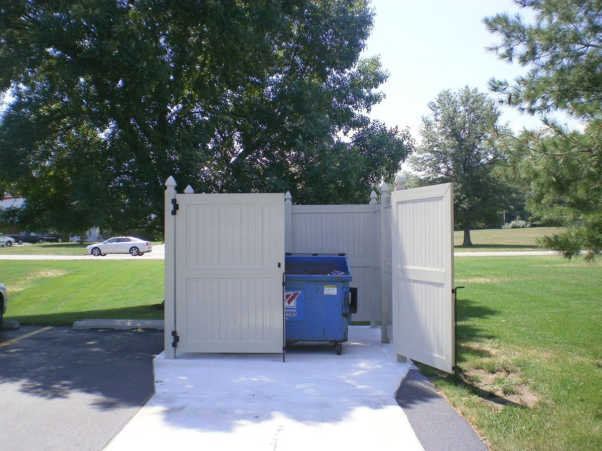 A blue dumpster inside a beige-fenced enclosure. The enclosure has two open doors, in a grassy area with a car.