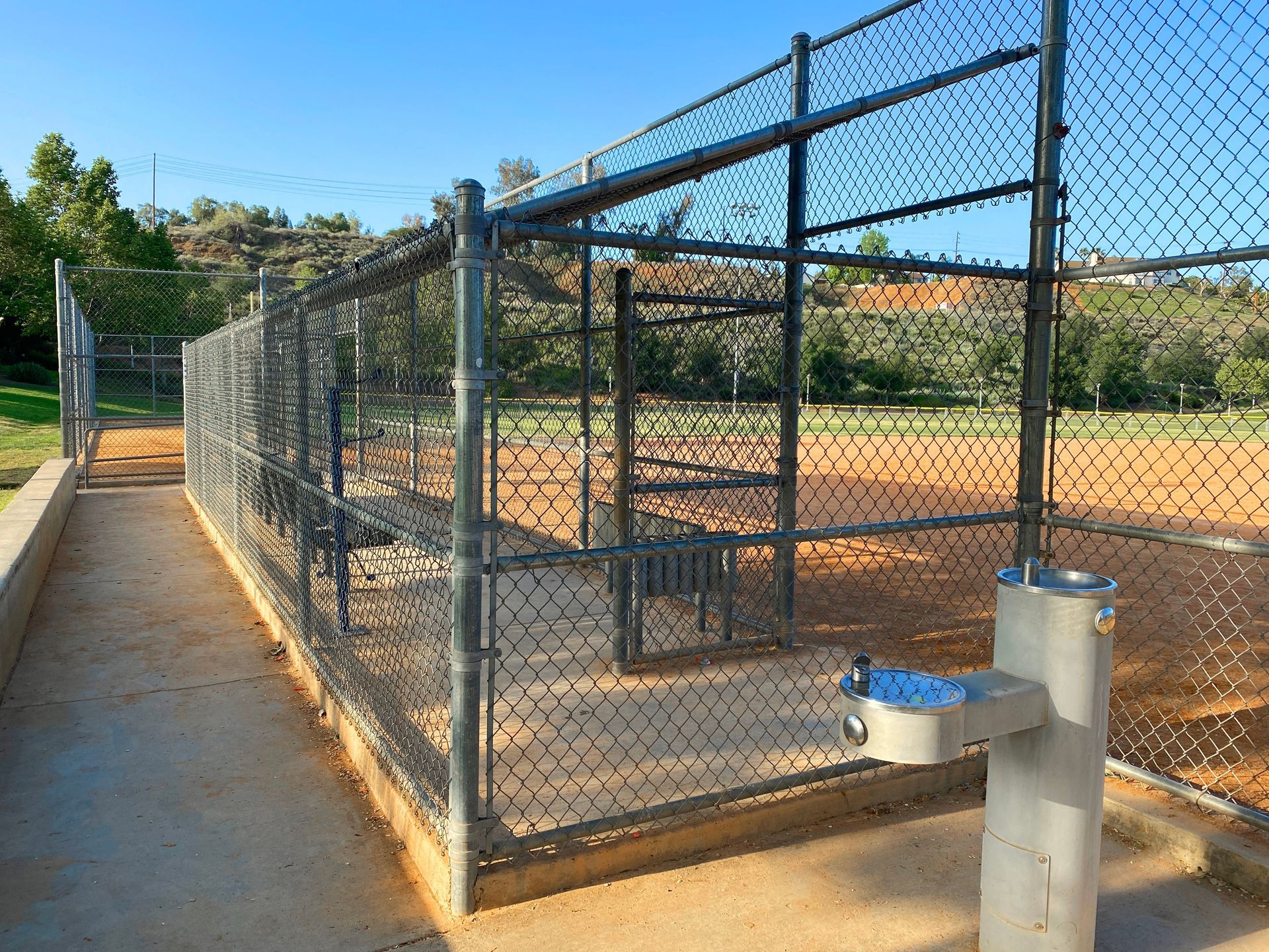 Dog park entrance with chain-link fence, benches, and a water fountain on a sunny day.