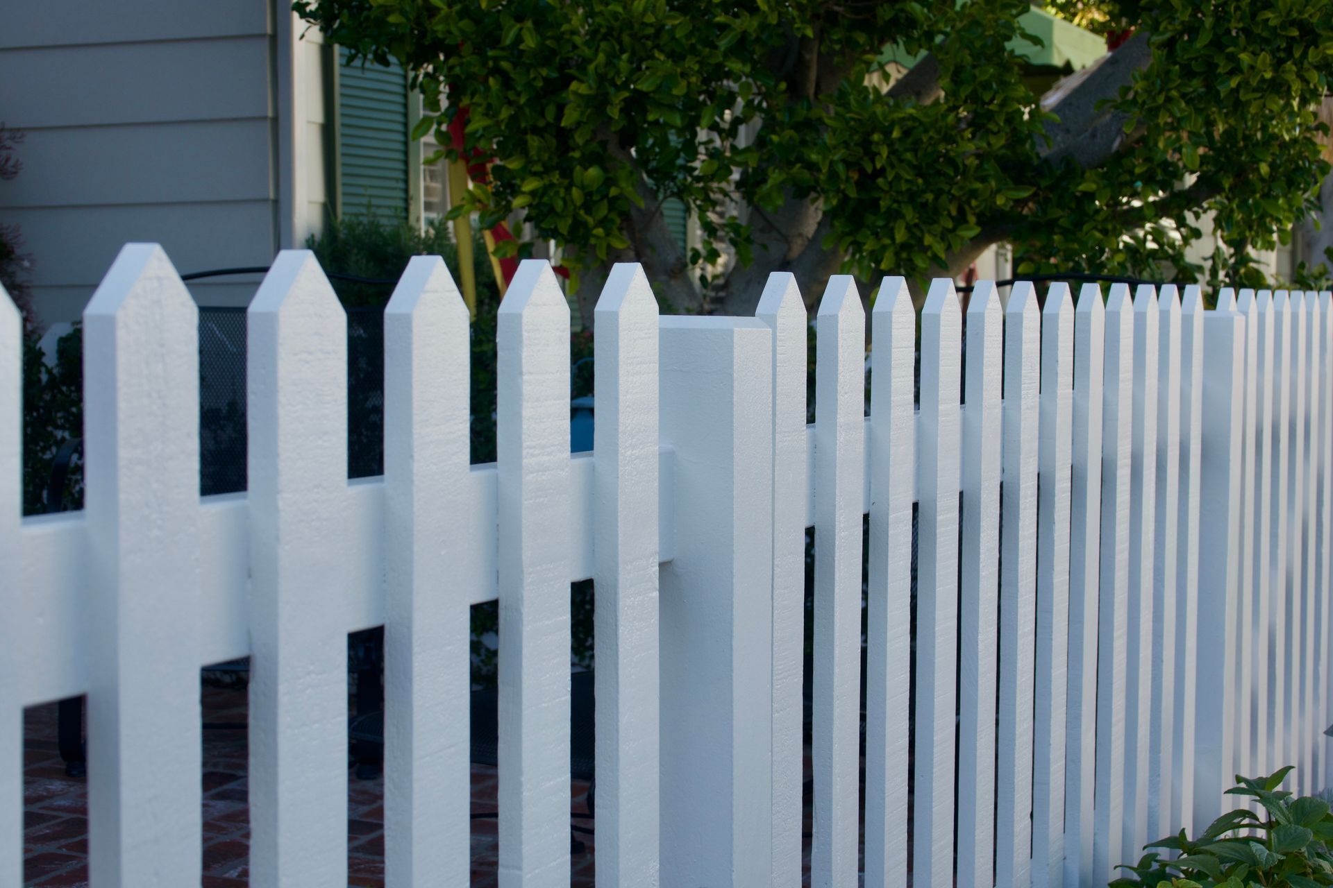White picket fence in front of a house, with green tree visible behind it.